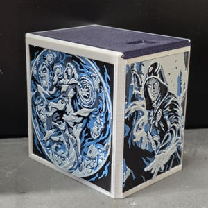 May include: A square storage box featuring 3D relief panels in shades of black, white, and light blue. The front panel displays a circular design with a central figure casting magic, while the side panel shows a hooded character in a dark cloak.