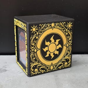 May include: A black rectangular card storage box featuring a gold-colored sun emblem surrounded by intricate filigree patterns on the side. The front frame is decorated with gold leaf-like motifs, and a single playing card is visible inside the box.