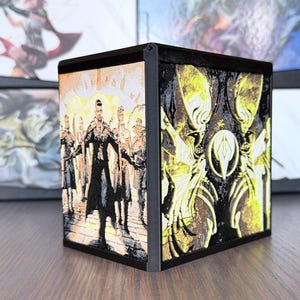 May include: A black, cube-shaped storage box with detailed artwork on its sides. One side features a group of figures in a city setting, while another displays a stylized, yellow-toned design. The box is likely for collectible cards or similar items.