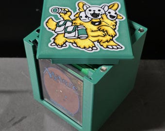 May include: A teal square deck box with a removable lid featuring a yellow three-headed creature illustration. The box has a clear front window displaying a Magic The Gathering card back. The container is constructed from a textured material.