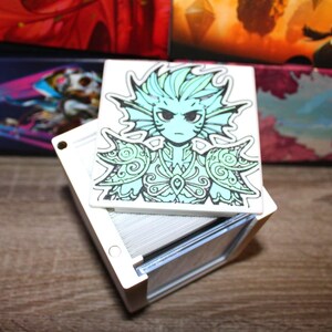 May include: A white card box with a stack of cards, topped with a square card featuring a teal and black illustration of a humanoid figure with aquatic features. The figure has a stern expression. Other boxes are in the background.