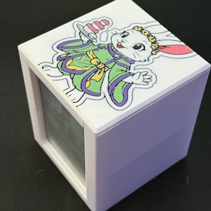 May include: A white, cube-shaped box with a hinged lid. The lid features a colorful illustration of a cartoon rabbit wearing a green and purple robe and a floral crown. The box has a clear window on one side.