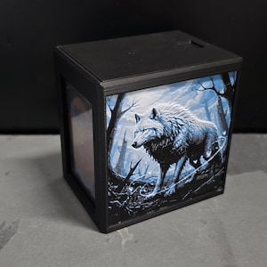 May include: Black box with a detailed illustration of a white wolf in a forest setting. The artwork features a wolf with blue and white tones, surrounded by bare trees. The box has a black frame.