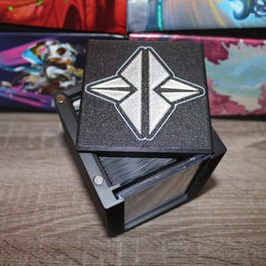 May include: Black card storage box with a silver geometric design on the lid. The box is open, revealing a stack of cards inside. The box is made of a textured material and has a hinged lid. Other boxes with colorful designs are in the background.