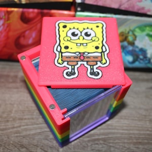May include: A pink card holder with a SpongeBob SquarePants design on the lid. The holder is stacked on a rainbow-colored base, with a clear plastic front. The holder is filled with blue cards.