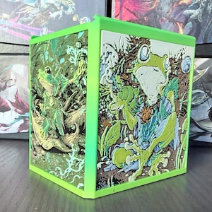 May include: A square, lime green container featuring relief-style panels on two visible sides. Each panel displays a detailed, multi-colored illustration of a frog sitting among foliage, rendered in shades of green, blue, and brown with textured surfaces.