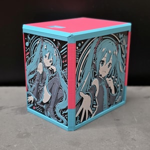 May include: A cubic storage box featuring a bright pink top and light blue frame. The side panels display a detailed relief illustration of a teal-haired anime character with headphones, set against a black background with musical notes and digital patterns.