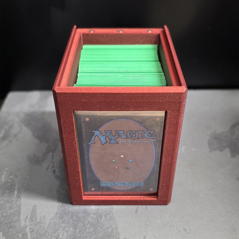 以下が含まれることがあります： 緑色のカードで満たされた赤いカード収納ボックス。ボックスには、Magic: The Gatheringカードの裏側が表示された透明な窓があります。ボックスは長方形で、テクスチャ加工された表面を持っています。
