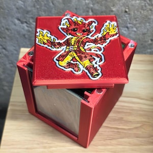 May include: A red, square box with a hinged lid. The lid features a colorful cartoon illustration of a devil-like character holding flames. The box is open, revealing a clear, reflective interior. The box is sitting on a wooden surface.