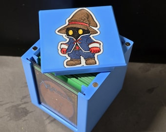 May include: A light blue square deck box with a removable lid featuring a graphic of a character wearing a brown pointed hat, blue coat, and red gloves. The box contains a stack of trading cards with green edges and a visible Magic The Gathering card back.