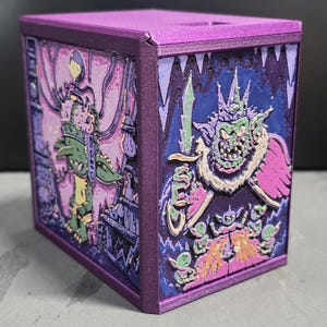 May include: A rectangular purple storage box featuring detailed relief panels on the sides. One panel depicts a green goblin-like figure with a sword, while the other shows a crowned character in a cave setting with smaller figures in shades of pink, blue, and green.
