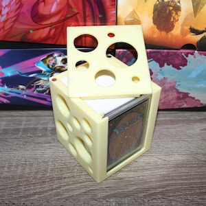 May include: A light yellow, cube-shaped card holder with circular cutouts and a removable top. The holder is designed to store trading cards, with a visible card partially inserted. The background includes colorful card storage boxes.