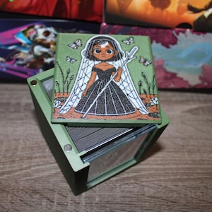 May include: A green box with a hinged lid, holding a deck of cards. The lid features a colorful illustration of a woman in a black dress, holding a staff, with butterflies and plants. Other boxes are visible in the background.