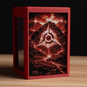 May include: A red card storage box with a striking design. The front panel features a fiery landscape with a white geometric symbol. The box is made of a solid material and has a clear window on the side.