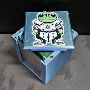 May include: A light blue 3D-printed deck box with a magnetic lid featuring a graphic of a green frog wearing a white and black knight's tunic with a star emblem. The box contains a stack of green-edged cards visible inside the open container.