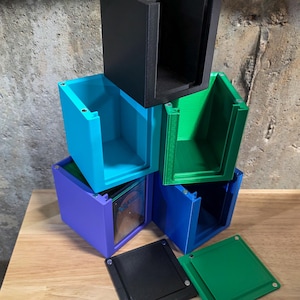May include: Stack of colorful, 3D-printed storage boxes in black, teal, green, blue, and purple. Each box has a rectangular shape with a lid. The boxes are stacked on a wooden surface against a textured gray wall.