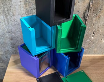 May include: Stack of colorful, 3D-printed storage boxes in black, teal, green, blue, and purple. Each box has a rectangular shape with a lid. The boxes are stacked on a wooden surface against a textured gray wall.