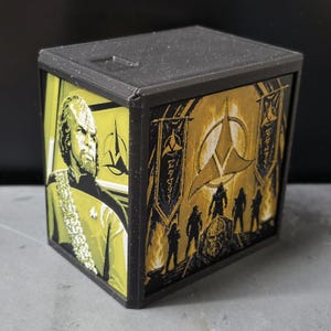 May include: A black rectangular storage box featuring two decorative panels with Star Trek Klingon imagery. One side displays a portrait of a Klingon character, while the other side shows a group of warriors with the Klingon Empire symbol.