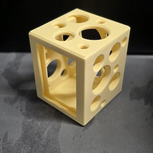 May include: A light yellow, cube-shaped decorative object with various sized circular cutouts. The object has a hollow interior and a solid base. The cube is made of a solid material and has a matte finish. The cutouts vary in size and shape.