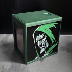 May include: A green box with a black front panel featuring a graphic of a character in a green hood. The box has a rectangular shape and a small rectangular opening on the top. The character graphic is in shades of green, white, and gray.