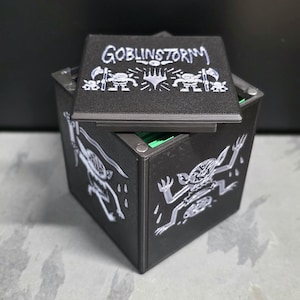 May include: A black square storage box with a removable lid featuring the text GOBLINSTORM and illustrations of goblins. The sides of the box are decorated with white line art of goblins, and the interior contains bright green dividers.