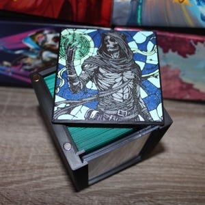 May include: A black card holder with a printed card featuring a skeletal figure in a hooded cloak, holding a glowing orb. The card has a blue and green stained-glass style background. The holder contains green cards.