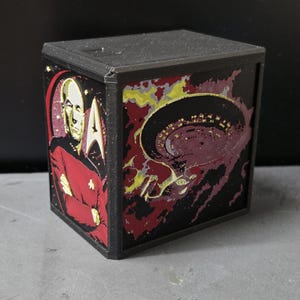 May include: A black 3D-printed cube featuring a relief image of Captain Jean-Luc Picard in a red uniform on one side and a depiction of the USS Enterprise starship against a dark, nebulous background on the adjacent side of the box.
