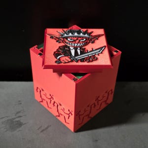 May include: A red square storage box with a removable lid featuring a graphic of a cartoon character wearing a spiked hat and suit while holding a serrated blade. The sides of the box are embossed with repeating patterns of small, stylized figures.