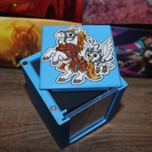 May include: A light blue square storage box with a removable lid. The lid features a colorful illustration of a white horse with a golden mane and a small white owl-like creature. The box has a dark interior and small silver magnets on the corners.