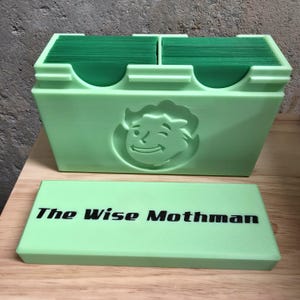 May include: A light green card holder with a winking face logo and the text "The Wise Mothman" on the lid. The holder is divided into two sections, each filled with green cards. The design is 3D printed.