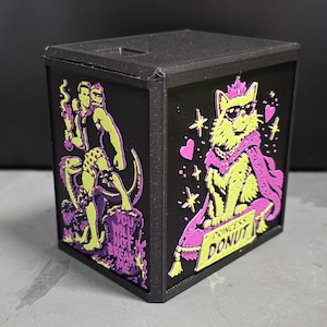 May include: A black cubic container featuring two distinct graphic panels. One side displays a figure with a dinosaur and the text "YOU WILL NOT BREAK ME." The other side shows a cat wearing a crown, sunglasses, and a cape with the text "PRINCESS DONUT."