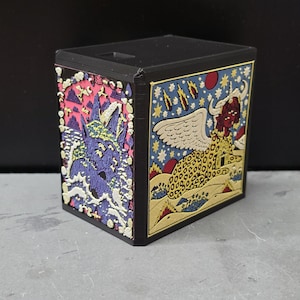 May include: A black rectangular box featuring two distinct relief panels. The left panel displays a purple creature in a pink sea with clouds. The right panel shows a winged, leopard-spotted creature with horns against a blue sky with falling stars.