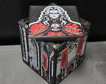 May include: A square black box with a lid featuring a graphic illustration of a long-haired character in a red cloak. The sides of the box are decorated with black and white architectural prints of gothic windows and red banners with symbols.