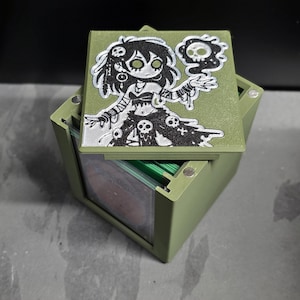 May include: A green card storage box with a hinged lid. The lid features a black and white illustration of a cartoon character with skulls. The box is open, revealing a stack of cards inside.