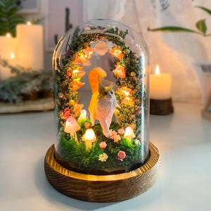 以下が含まれることがあります： 猫の置物、光るキノコ、花のアクセントを特徴とするミニチュアシーンを備えた装飾的なガラスドーム。ベースは木製です。シーンは暖かく柔らかな光で照らされ、気まぐれで魅惑的なディスプレイを作り出しています。