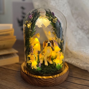 Peut inclure: Un dôme en verre décoratif avec une figurine de beagle, des plantes artificielles, des champignons lumineux et des guirlandes lumineuses. Le dôme est posé sur une base en bois, créant un affichage lumineux. Le beagle a des marques marron et blanches.