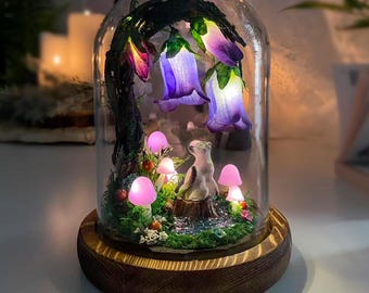 Puede incluir: Una cúpula de cristal decorativa con una escena de bosque. En su interior, hay flores de campana moradas brillantes, setas rosas y una pequeña figura de animal. La cúpula se asienta sobre una base de madera, creando una exhibición caprichosa e iluminada.
