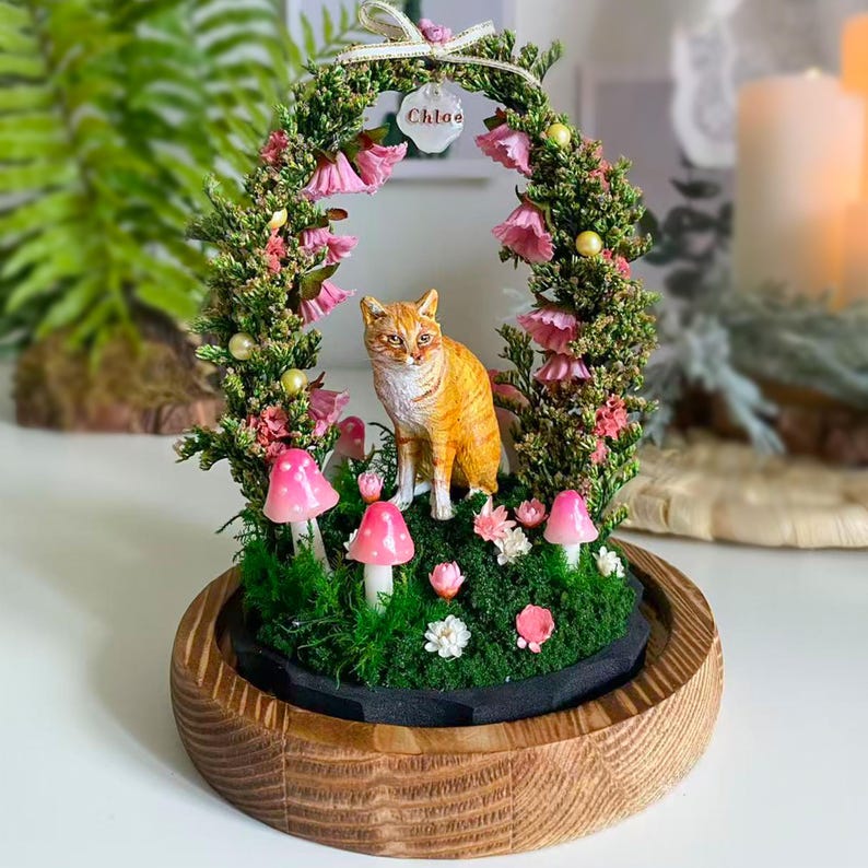 以下が含まれることがあります： ピンクのキノコ、花、緑に囲まれた、オレンジ色の猫の置物が入った装飾的なガラスドーム。猫は木製の台座の中の苔の上に座っています。「Chloe」という名前のフローラルアーチが猫の上にあります。