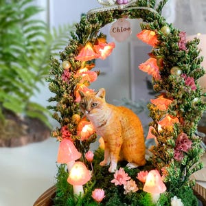 以下が含まれることがあります： オレンジ色の猫の置物がミニチュアガーデンに置かれています。庭には光るキノコのライト、ピンクの花、そして「Chloe」という名前のタグが付いた花のアーチがあります。全体的なデザインは気まぐれで詳細です。
