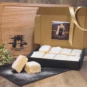 May include: A box of shortbread cookies dusted with powdered sugar, with two cookies in front. The box has a Sugar Pony label with a horse image. A wooden cutting board with a Sugar Pony logo is in the background.