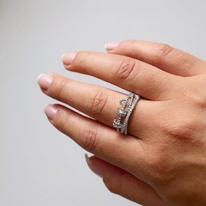 Puede incluir: Un anillo de corona plateado con pequeñas piedras preciosas transparentes. El anillo, que se lleva en un dedo, tiene un diseño de corona con múltiples capas y elementos decorativos. El fondo es neutro.