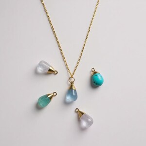 Può includere: Una collana dorata con un ciondolo a forma di goccia di pietra blu. Quattro pietre preziose aggiuntive in vari colori, tra cui trasparente, turchese e verde, sono sparse intorno alla collana. Ogni pietra ha un cappuccio dorato.