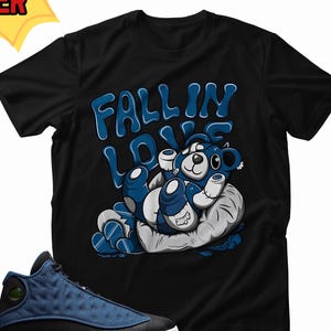 Pode incluir: Camiseta preta com um urso de desenho animado azul e branco e o texto "FALL IN LOVE". Um tênis azul e preto com solas brancas está em primeiro plano.