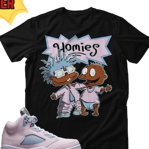 May include: Black t-shirt with a cartoon graphic of two characters and the word "Homies" in pink and blue. A pair of pink and light blue sneakers are in the foreground.
