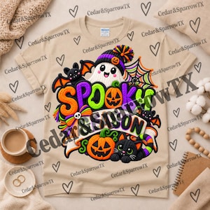 May include: A beige t-shirt featuring a colorful graphic with the text SPOOKY SEASON. The design includes a white ghost wearing a purple and orange pom-pom hat, orange pumpkins, a black cat, a black bat, a spider web, and a small skull.