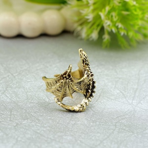 Könnte beinhalten: Ein goldfarbener Drachenring mit aufwendigen Details. Der Ring zeigt einen Drachen mit ausgebreiteten Flügeln und einem strukturierten Körper. Der Schwanz des Drachen umschlingt das Ringband und schafft ein einzigartiges Fantasy-Accessoire.