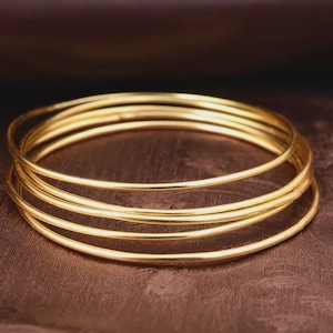 Può includere: Una pila di cinque braccialetti d'oro, ciascuno con una superficie liscia e arrotondata. I braccialetti sono disposti in modo nidificato, creando un effetto a strati. I gioielli sono su uno sfondo marrone scuro strutturato.