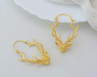 May include: A pair of gold-colored hoop earrings. Each earring features a deer head design with detailed antlers. The earrings are displayed on a white surface, showcasing their intricate craftsmanship and elegant style.