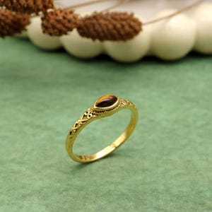 Pode incluir: Um anel dourado com uma pedra preciosa oval marrom. O anel tem um design ornamentado de filigrana ao longo da banda e ao redor da configuração da pedra preciosa. O anel está sobre um fundo verde.