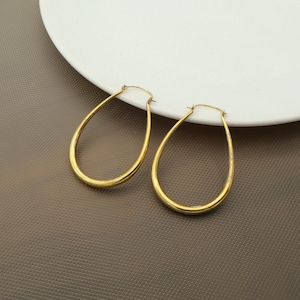 Può includere: Un paio di orecchini a cerchio dorati a forma di goccia. Gli orecchini hanno una finitura liscia e lucida e un design classico ed elegante. Sono esposti su un piatto bianco su sfondo marrone.